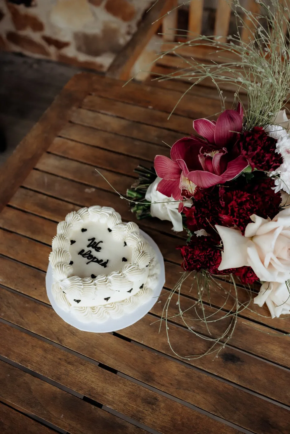 A small white heart-shaped cake beside burgundy and cream flowers by The Romantics on a timber table. At Piermont Retreat on Tasmania’s east coast, for an Elopement Collective elopement. Photograph by Omar of By The Wilde.