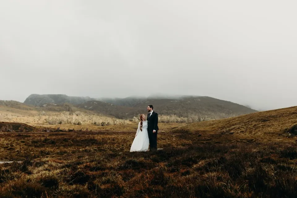 A couple stand among golden alpine grasses with mist-covered mountains behind them.