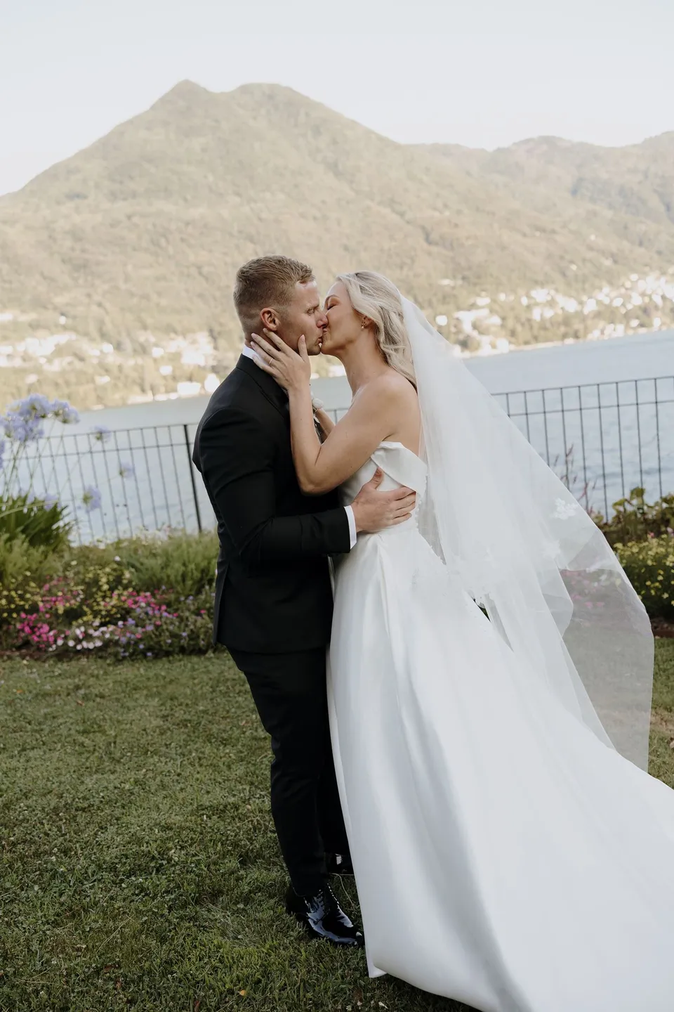 Zoe and Charlton kissing on the ceremony lawn, with her veil falling over the grass and the lake behind them, at Passalacqua in Moltrasio, Lake Como, Italy, for an Elopement Collective elopement. Photo by George Bowden.