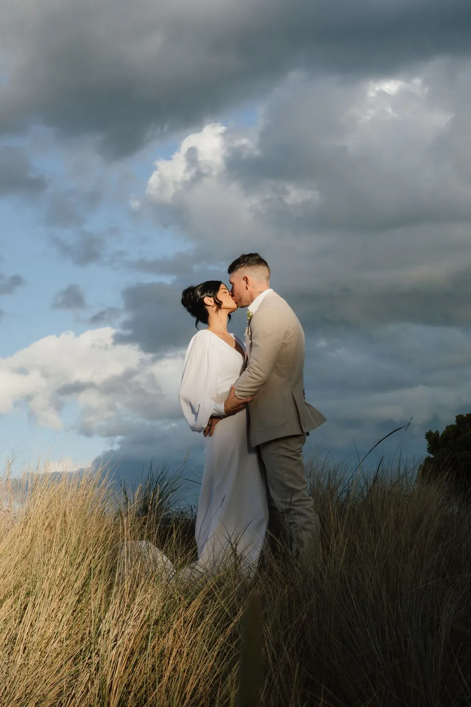 Renee and Mitch kissing among sunlit coastal grasses beneath dramatic clouds above the bay. At Piermont Retreat on Tasmania’s east coast, for an Elopement Collective elopement. Photograph by Bulb Creative.