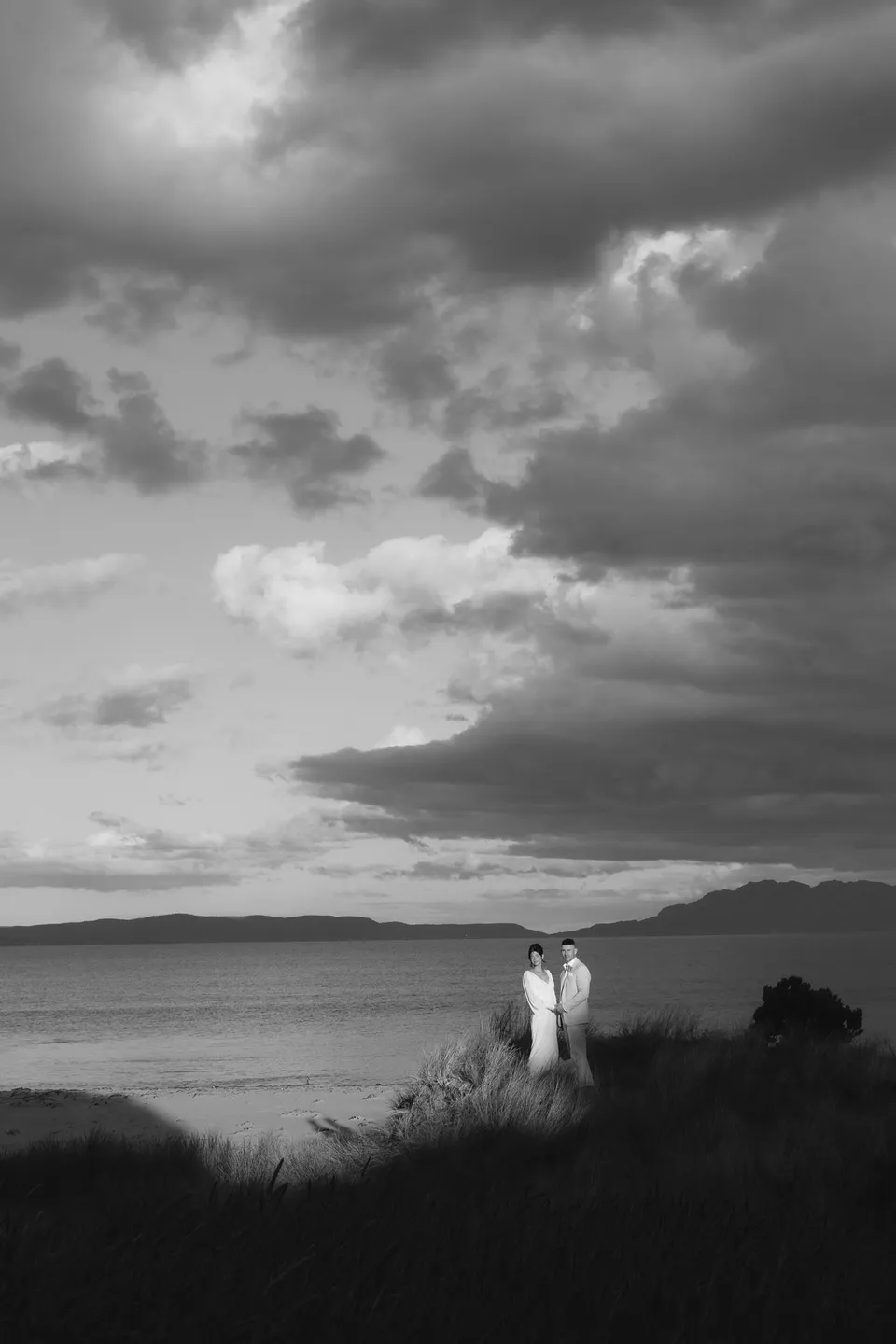 Black and white photograph of Renee and Mitch standing above the shoreline beneath large clouds over the bay. At Piermont Retreat on Tasmania’s east coast, for an Elopement Collective elopement. Photograph by Bulb Creative.