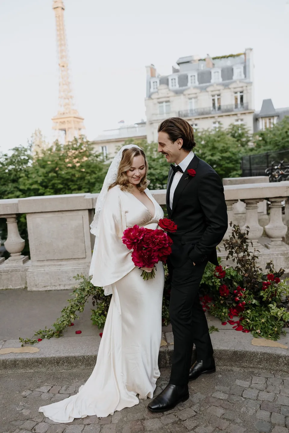 Kara and Samuel standing together beside the ceremony setup, with her bouquet and his red buttonhole by Ivy Bleu and the Eiffel Tower beyond, at Avenue de Camoëns in Paris, France, during an Elopement Collective elopement. Photograph by George Bowden.