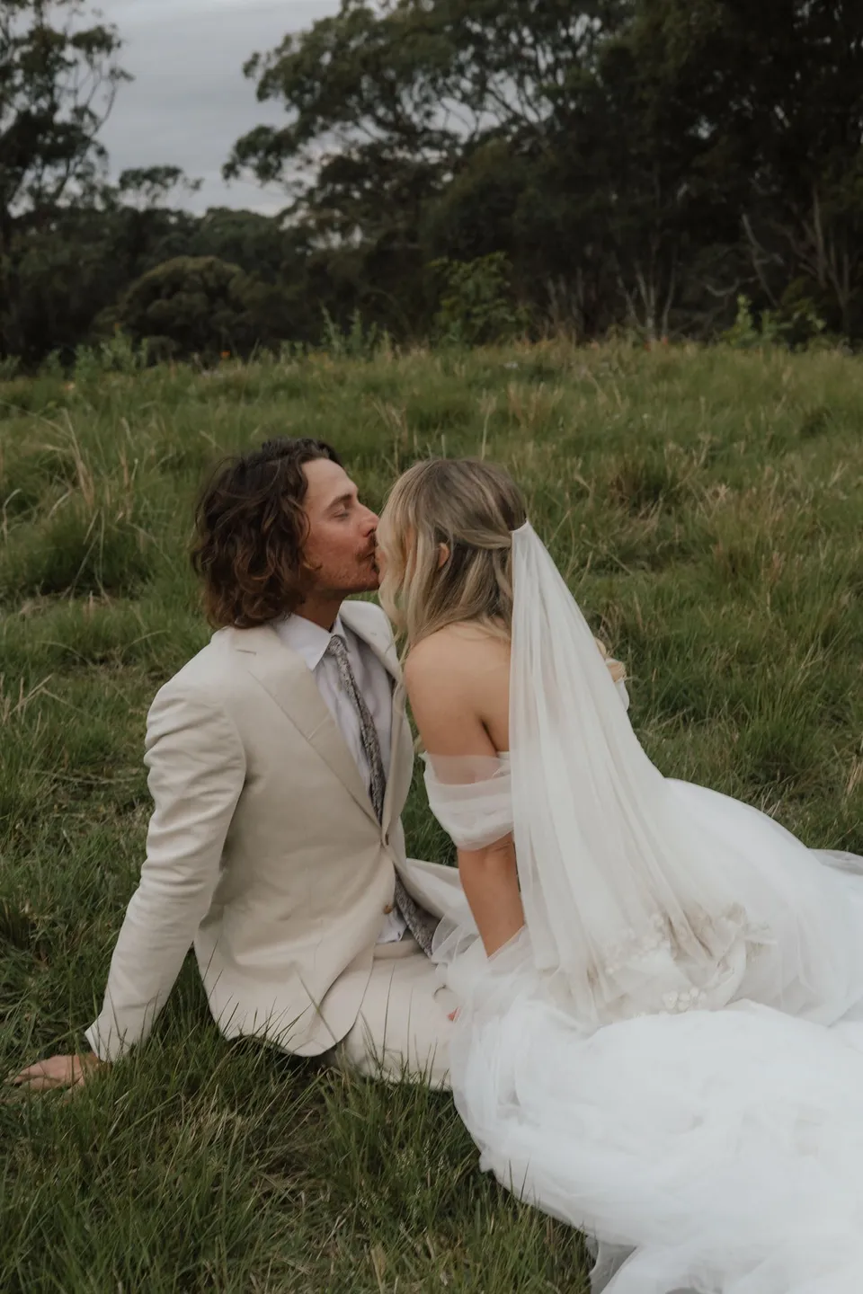 Bridget and James kissing while seated together in the long grass. At Cudgera Creek in northern New South Wales, for an Elopement Collective elopement. Photograph by Bulb Creative.