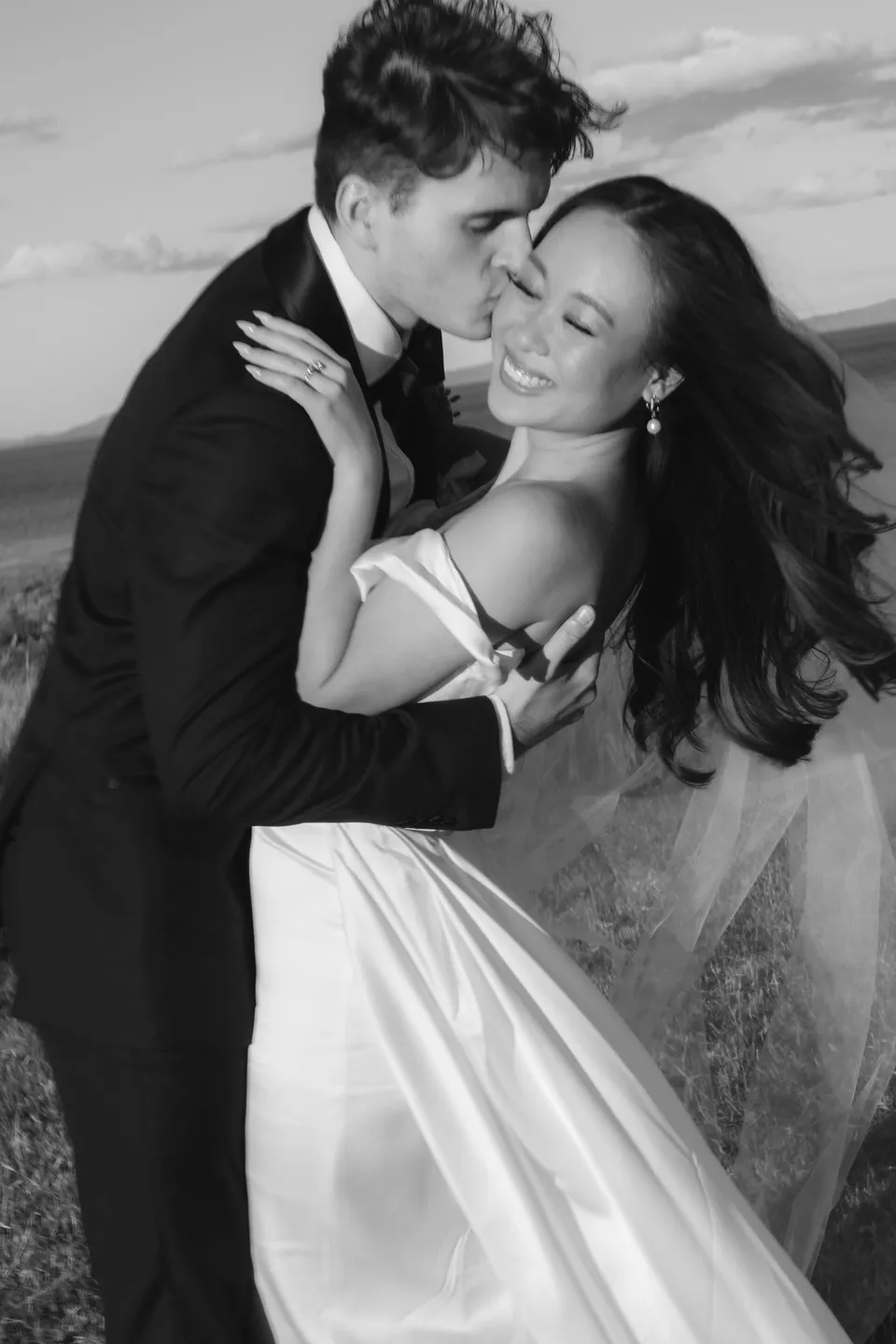 Black and white portrait of Harry kissing Clara’s cheek as she laughs and embraces him, at Thalia Haven on the East Coast of Tasmania, for an Elopement Collective elopement. Photograph by Bulb Creative.