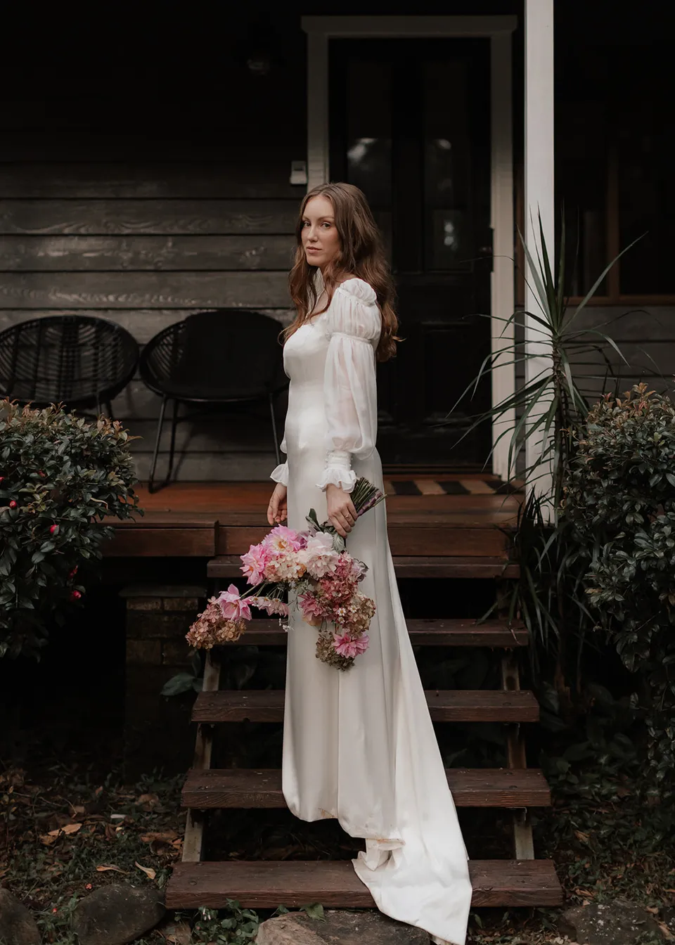 Elyse standing beside the cottage steps with her bouquet, with flowers by The Perfect Party Co. At Fig Tree Villa in Ewingsdale, Byron Bay, for an Elopement Collective elopement. Photograph by Bulb Creative.