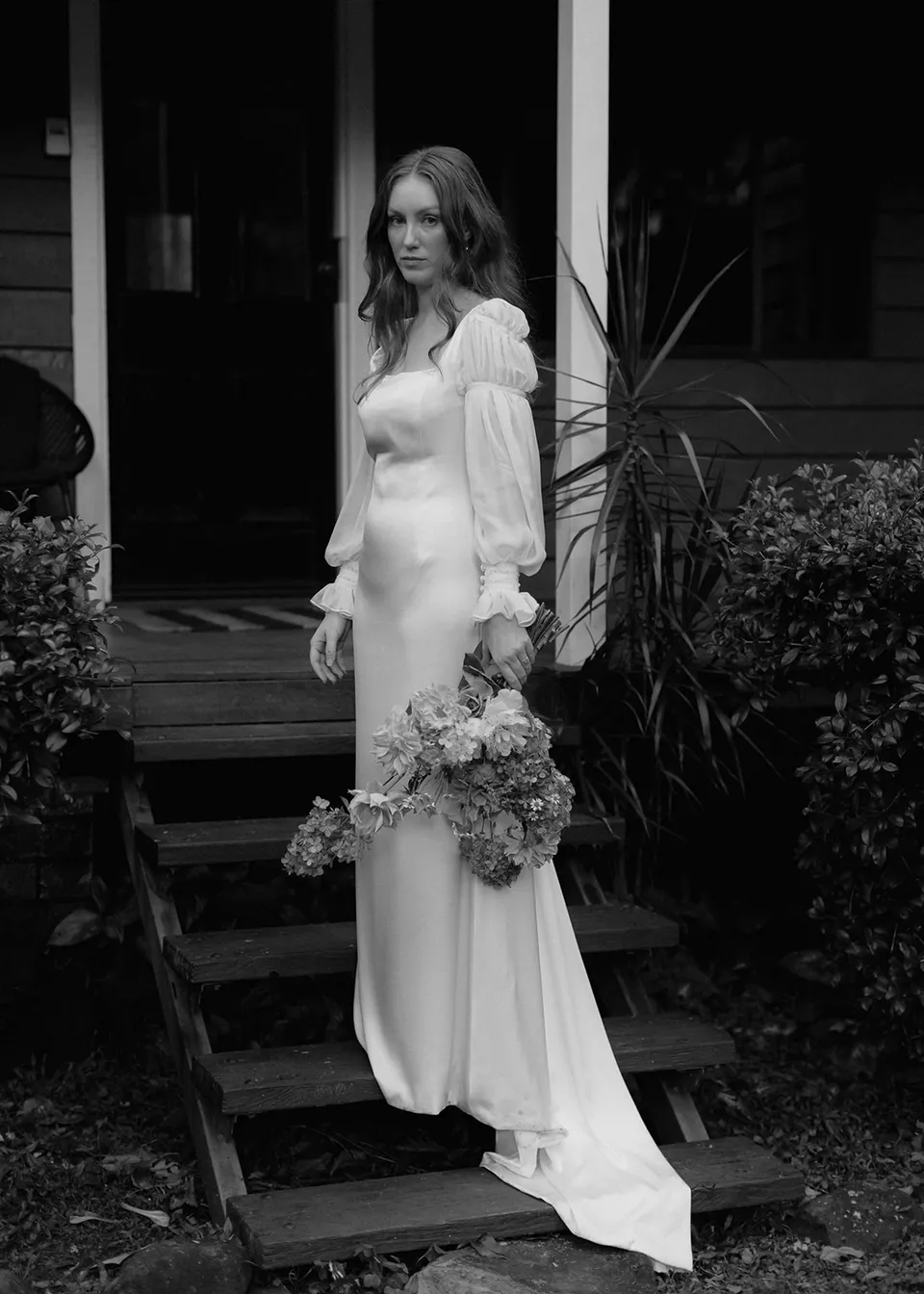 Black-and-white portrait of Elyse at the foot of the cottage steps. At Fig Tree Villa in Ewingsdale, Byron Bay, for an Elopement Collective elopement. Photograph by Bulb Creative.