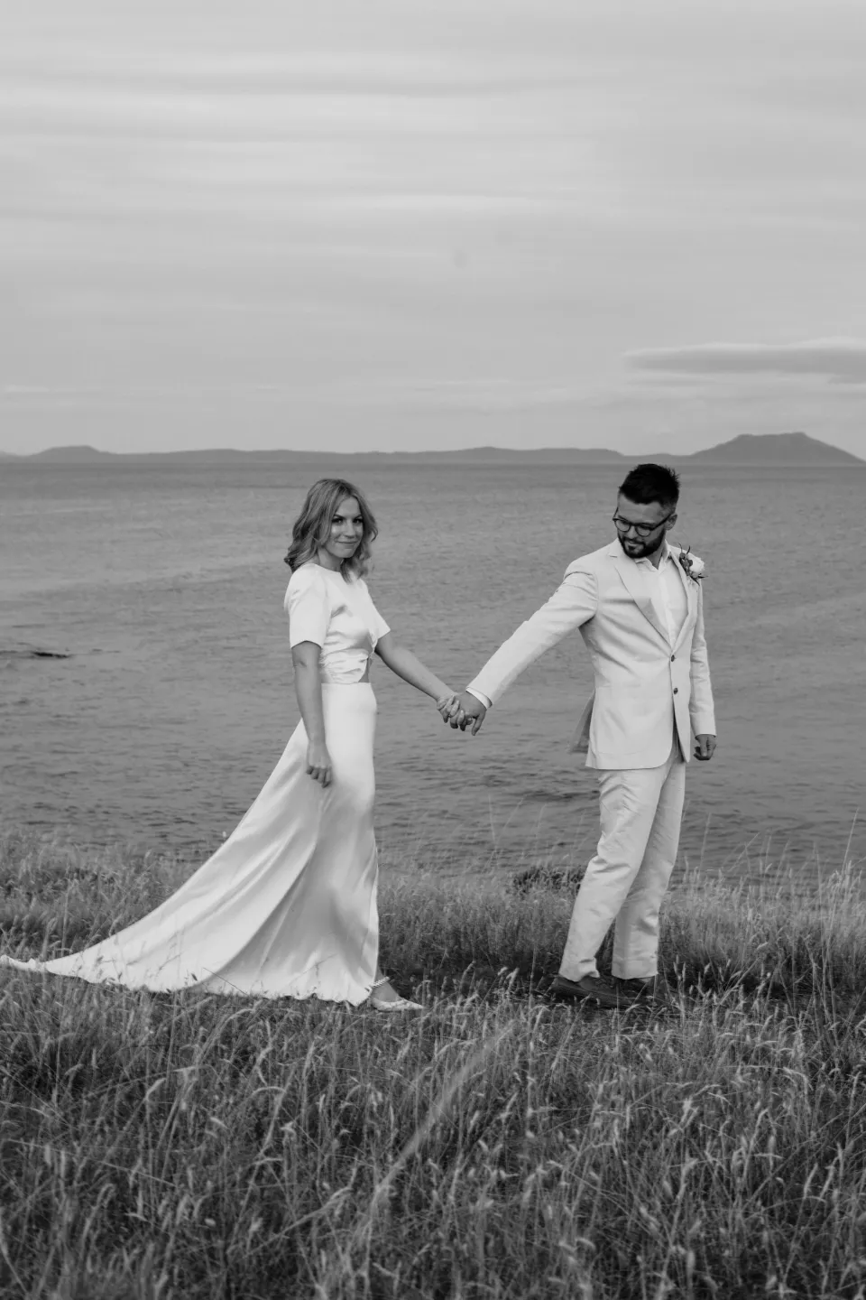 Black and white photograph of Jemmah and Jack holding hands in the meadow, at Thalia Haven on the East Coast of Tasmania, Australia, for an Elopement Collective elopement. Photograph by Joey Willis.
