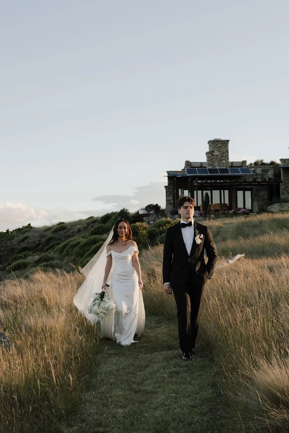Clara and Harry walking towards the camera along a narrow grassy path, with her white bouquet by The Romantics and the stone house behind them, at Thalia Haven on the East Coast of Tasmania, for an Elopement Collective elopement. Photograph by Bulb Creative.