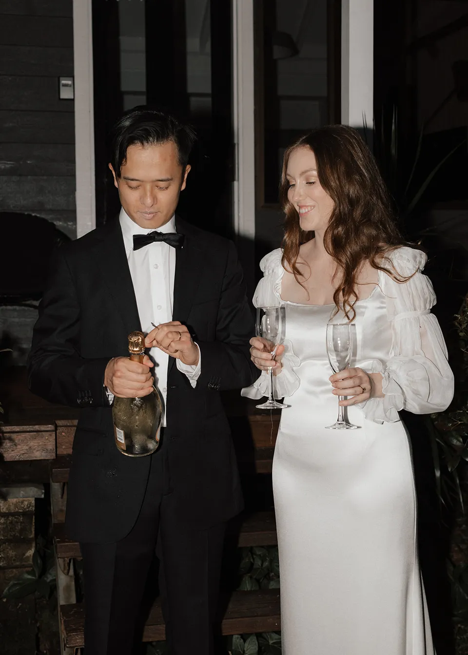 Jonathan opening sparkling wine while Elyse holds their glasses. At Fig Tree Villa in Ewingsdale, Byron Bay, for an Elopement Collective elopement. Photograph by Bulb Creative.