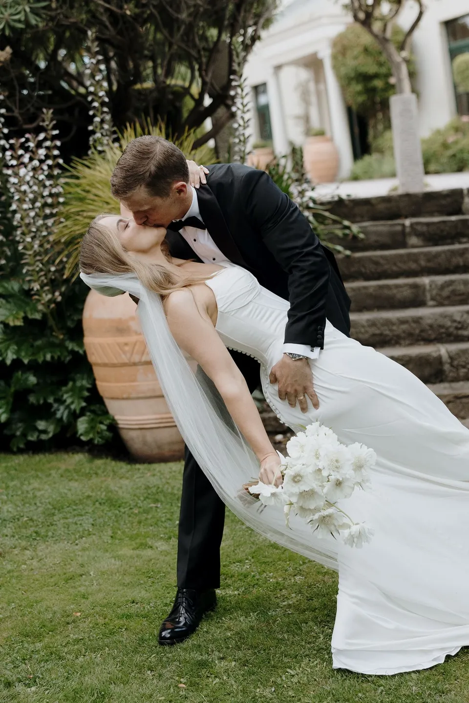 Andrew dipping Tanesha into a kiss beside the garden steps, with her white bouquet by The Romantics and veil falling towards the lawn, at Islington Hotel in South Hobart, Tasmania, Australia, for an Elopement Collective summer elopement. Photograph by George Bowden.