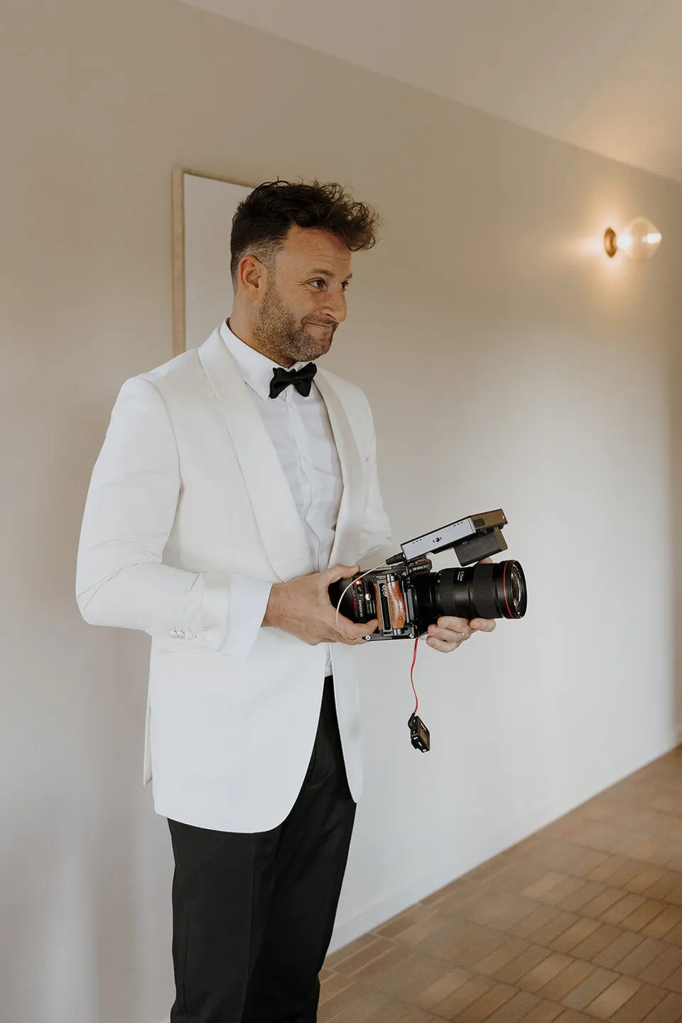 Greg holding a camera inside the cabin, wearing a white dinner jacket and black bow tie, at Roundhill Retreat in Obi Obi, Sunshine Coast Hinterland, Queensland, for an Elopement Collective elopement. Photo by George Bowden.