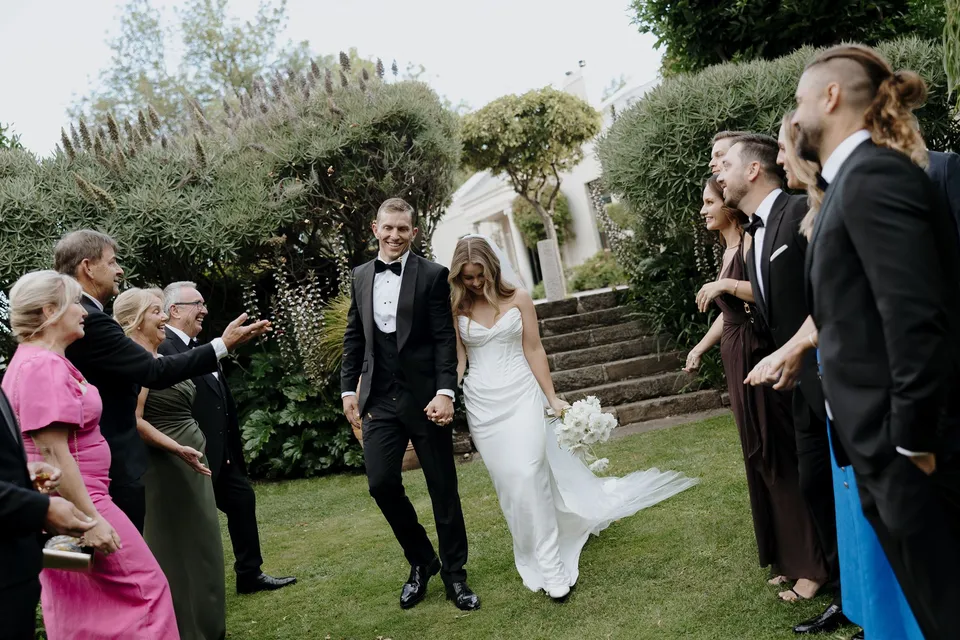 Tanesha and Andrew walking hand in hand between smiling guests after their ceremony, with her bouquet by The Romantics at her side, at Islington Hotel in South Hobart, Tasmania, Australia, for an Elopement Collective summer elopement. Photograph by George Bowden.