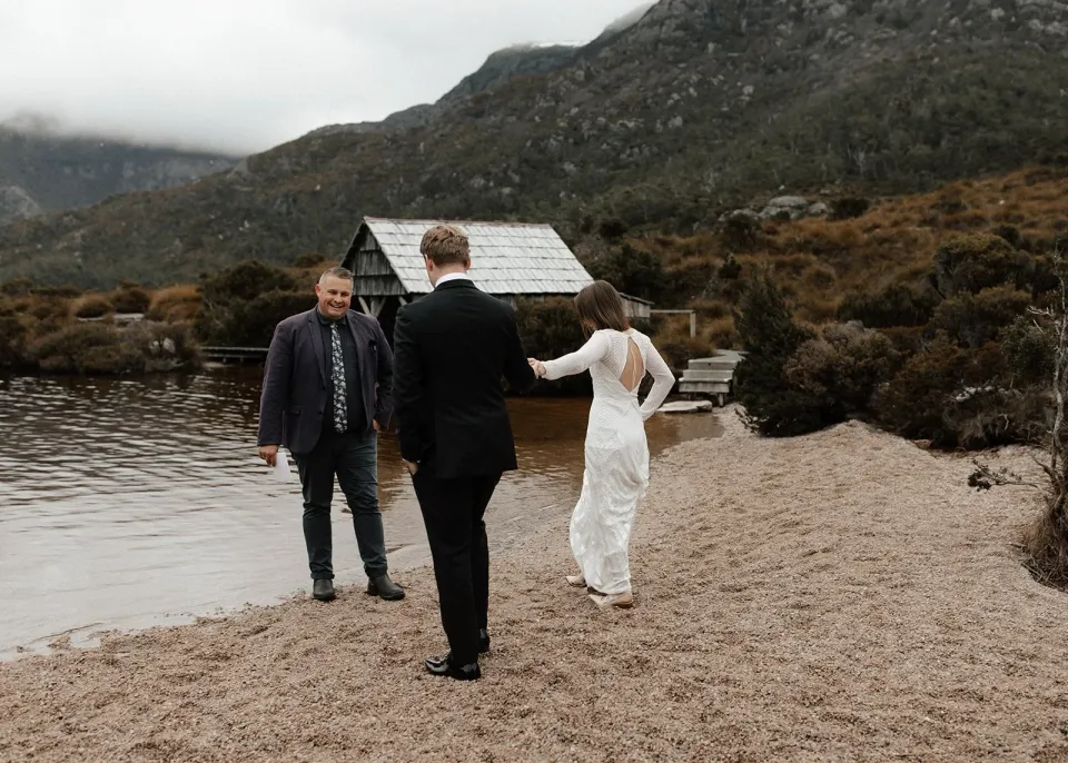 A couple and their celebrant walk beside a mountain lake and a small timber boatshed.