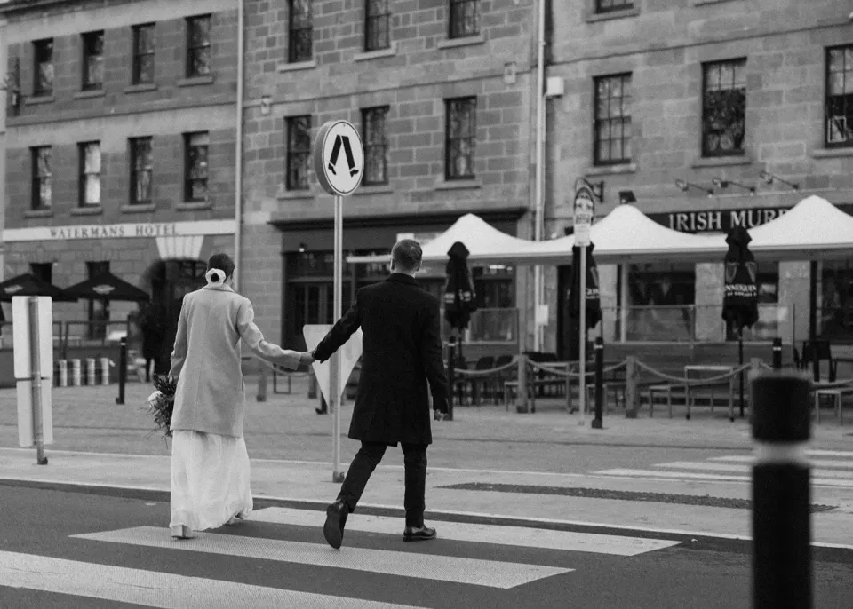 A black-and-white photograph of a couple holding hands across a street lined with sandstone buildings.