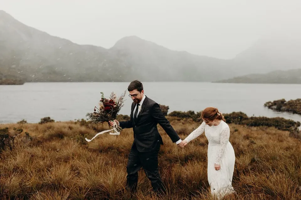 A couple walk hand in hand through tussock grass beside a misty lake.