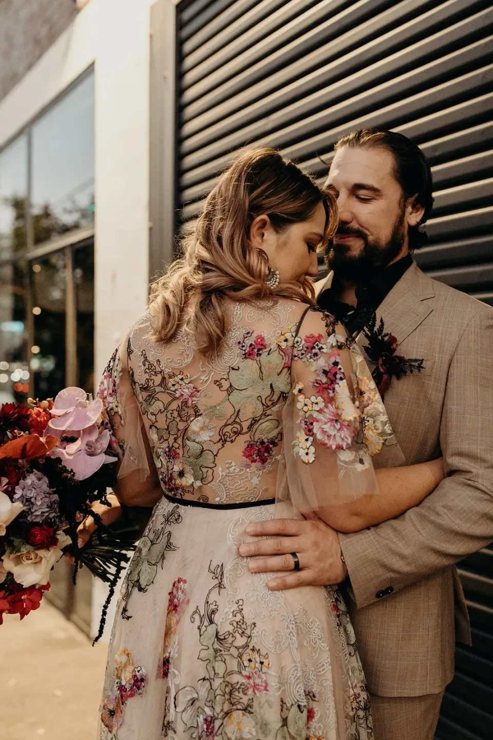 Victoria and Daniel embracing beside a black roller door, with her bouquet by Mrs Gibbons Flowers, in Fortitude Valley, Brisbane, for an Elopement Collective elopement. Photo by Tessa Shannon.