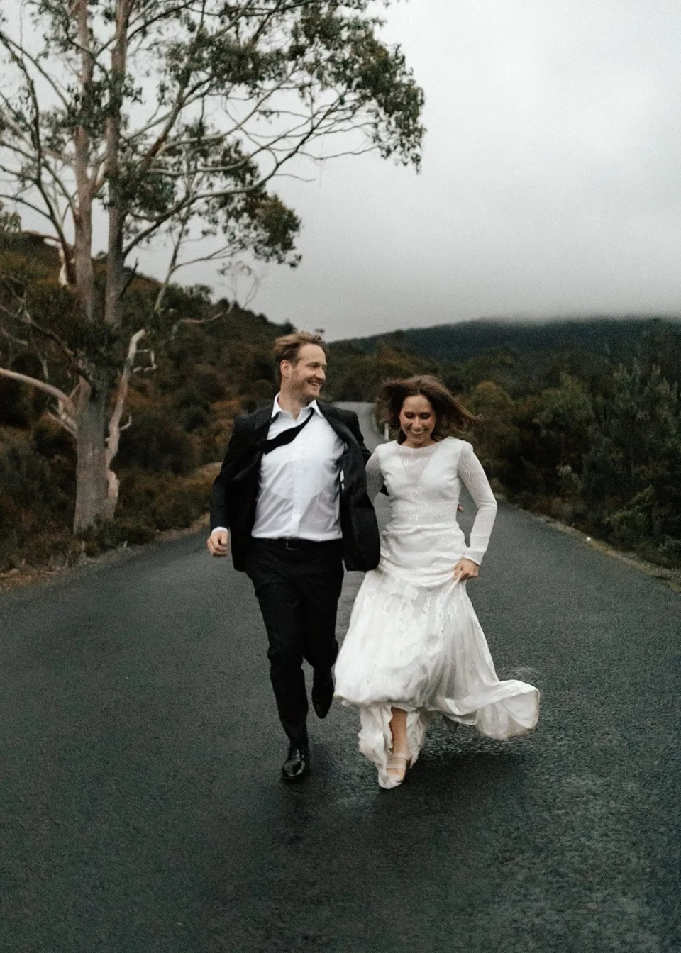 April and Josh running hand in hand along a wet road, her white dress lifting as they move, in Cradle Mountain, Tasmania, for an Elopement Collective elopement. Photo by Bulb Creative.