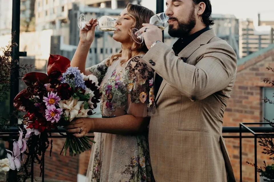 Victoria and Daniel drinking sparkling wine after their rooftop ceremony, with her bouquet by Mrs Gibbons Flowers, at Agnes Restaurant in Fortitude Valley, Brisbane, for an Elopement Collective elopement. Photo by Tessa Shannon.