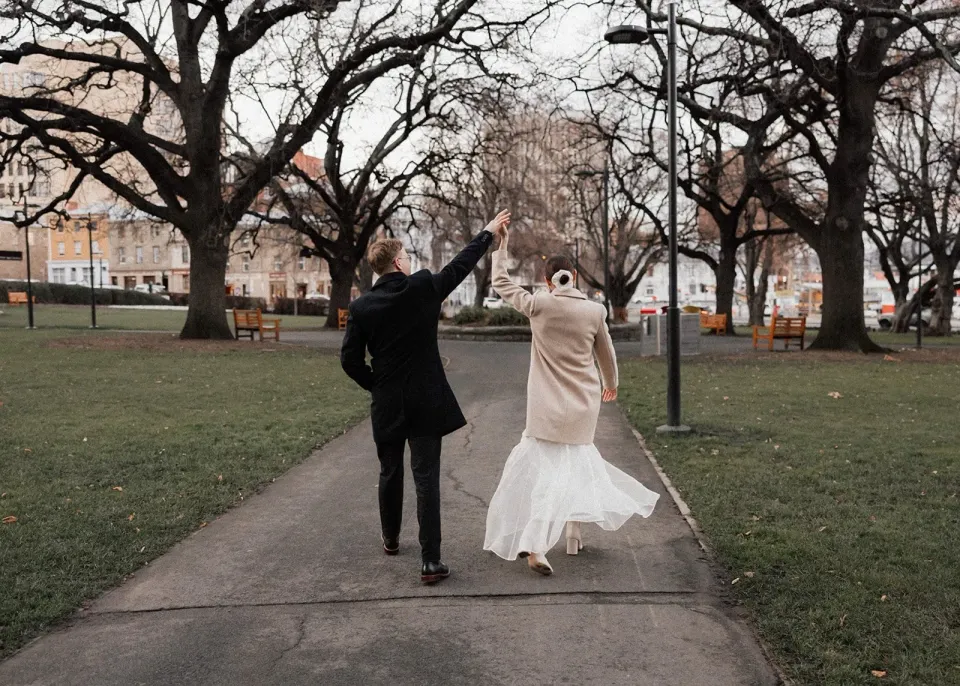 A groom twirls his bride along a garden path lined with bare trees.