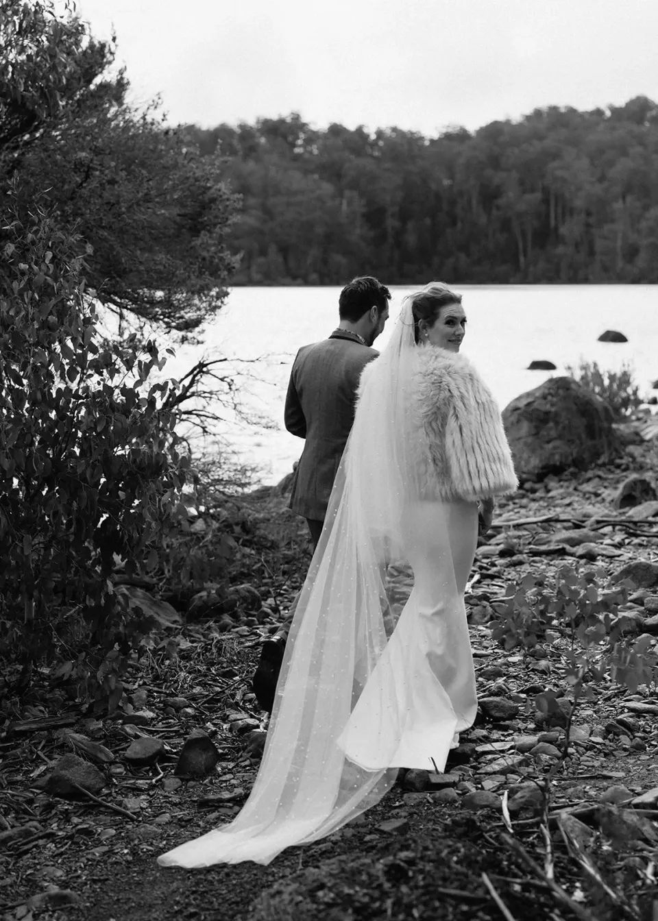 Black-and-white portrait of Sarah looking back over her shoulder while walking beside Jono on the lakeshore, at Pumphouse Point, Lake St Clair, Tasmania, for an Elopement Collective elopement. Photo by Bulb Creative.