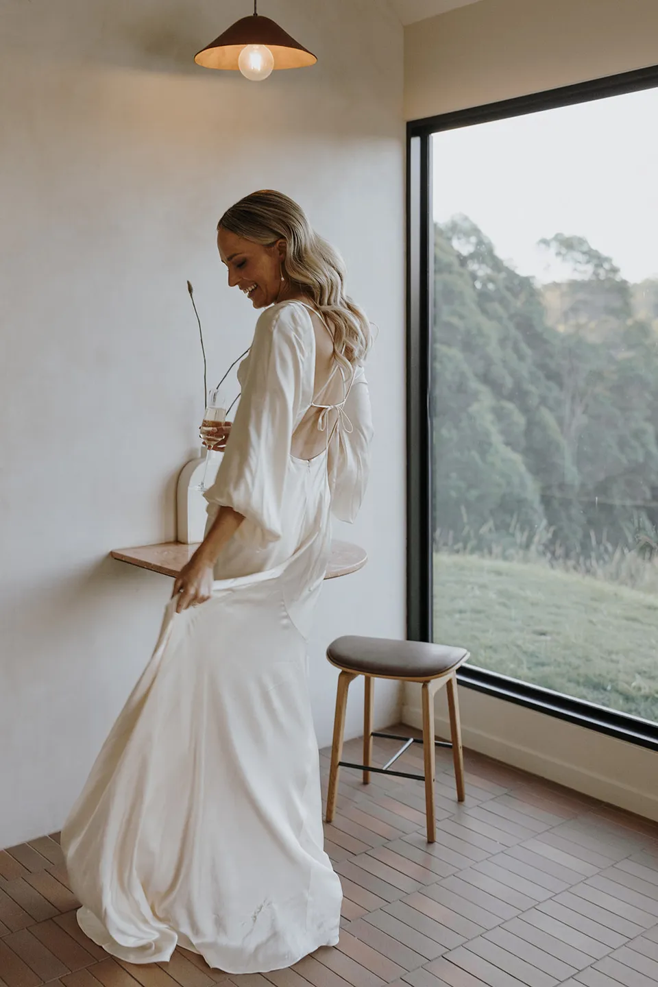 Gabrielle standing beside the cabin’s large window, her ivory Shona Joy dress trailing across the tiled floor, at Roundhill Retreat in Obi Obi, Sunshine Coast Hinterland, Queensland, for an Elopement Collective elopement. Photo by George Bowden.