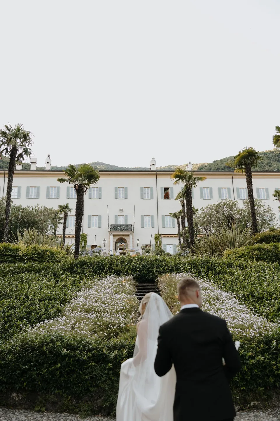Zoe and Charlton walking away along a path through clipped hedges towards Passalacqua’s pale villa, at Passalacqua in Moltrasio, Lake Como, Italy, for an Elopement Collective elopement. Photo by George Bowden.