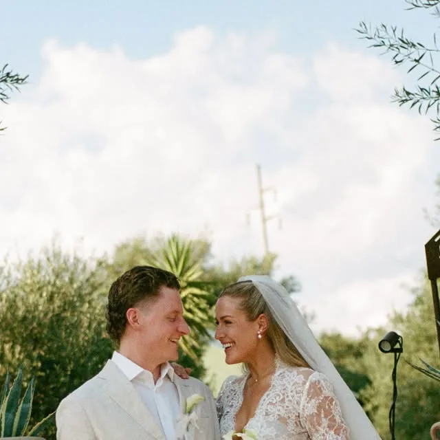 A couple smiling at each other outdoors in wedding clothes.