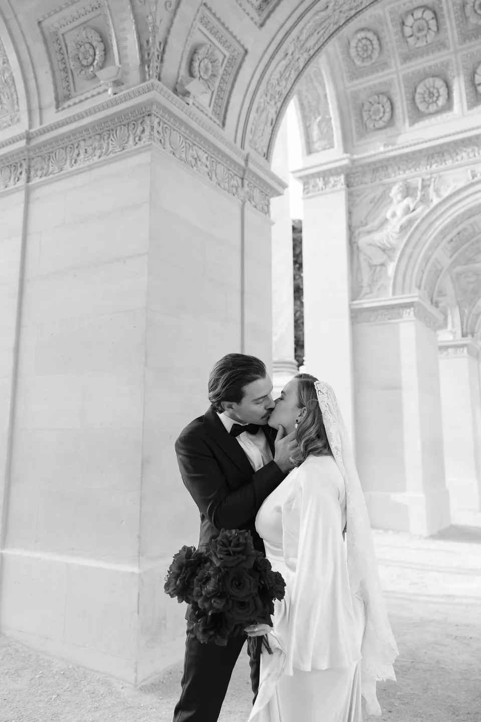 Black and white photograph of Kara and Samuel embracing beneath an arched stone colonnade, at the Louvre in Paris, France, during an Elopement Collective elopement. Photograph by George Bowden.