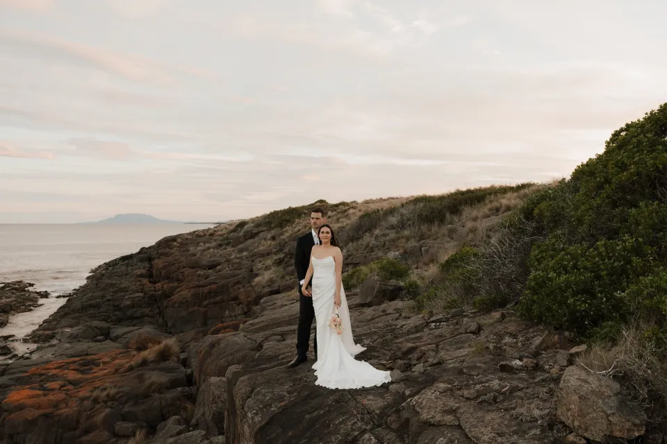 Dani and Anthony standing on a broad coastal rock ledge, with her white dress, shoreline plants and the bay around them, at Thalia Haven on Tasmania’s east coast, for an Elopement Collective elopement. Photograph by Bulb Creative.