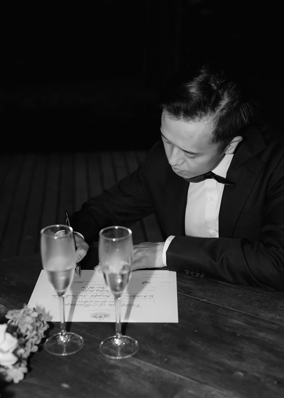 Black-and-white photograph of Jonathan signing paperwork beside two glasses. At Fig Tree Villa in Ewingsdale, Byron Bay, for an Elopement Collective elopement. Photograph by Bulb Creative.