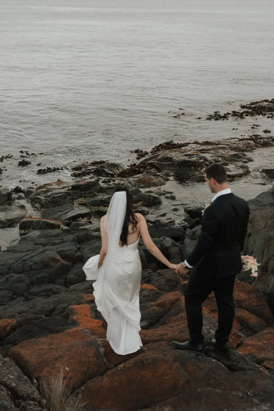 Dani and Anthony holding hands as they make their way down the layered rocks towards the water, at Thalia Haven on Tasmania’s east coast, for an Elopement Collective elopement. Photograph by Bulb Creative.