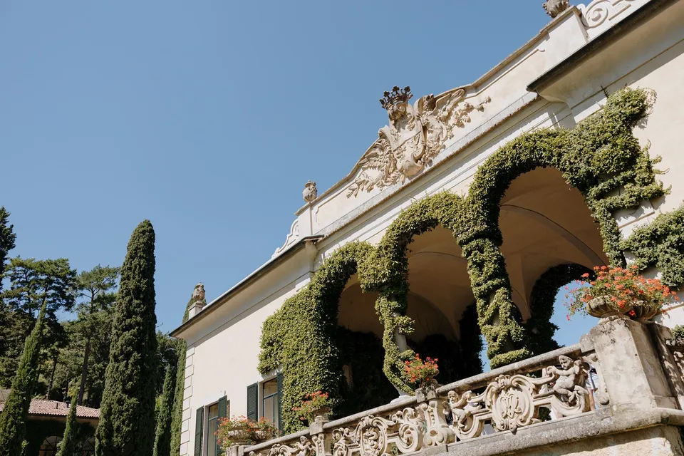 Low view of Villa del Balbianello’s ivy-covered loggia, carved stone balustrade and cypress trees beneath a blue sky, at Lake Como in Lombardy, Italy, for an Elopement Collective elopement. Photograph by George Bowden.