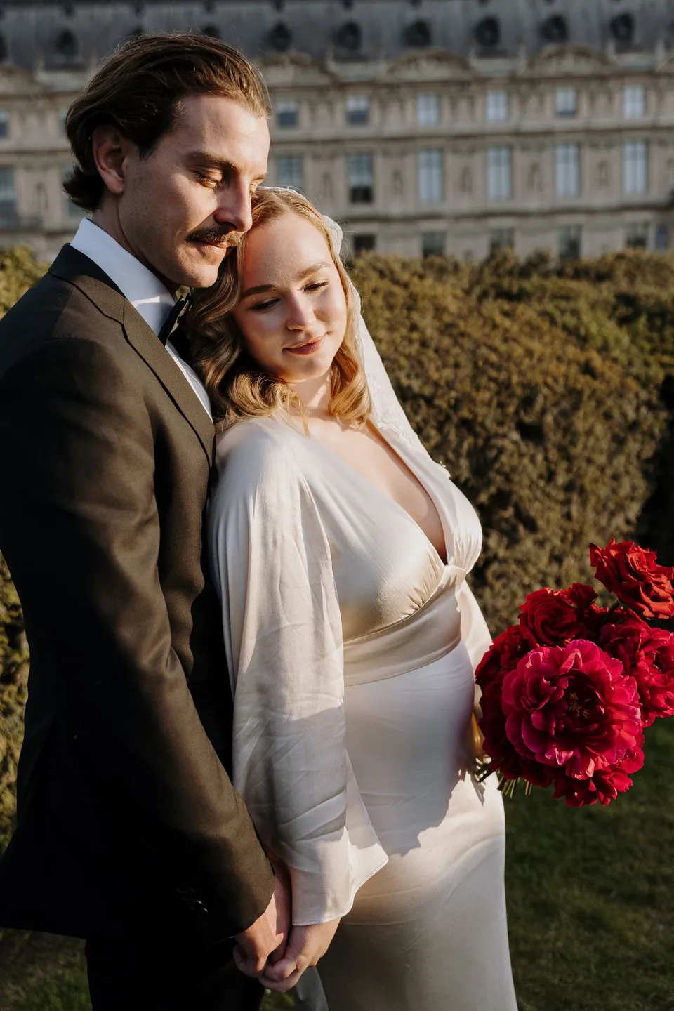 Kara and Samuel standing close in warm light beside a hedge and palace facade, with her red Ivy Bleu bouquet, at the Louvre in Paris, France, during an Elopement Collective elopement. Photograph by George Bowden.