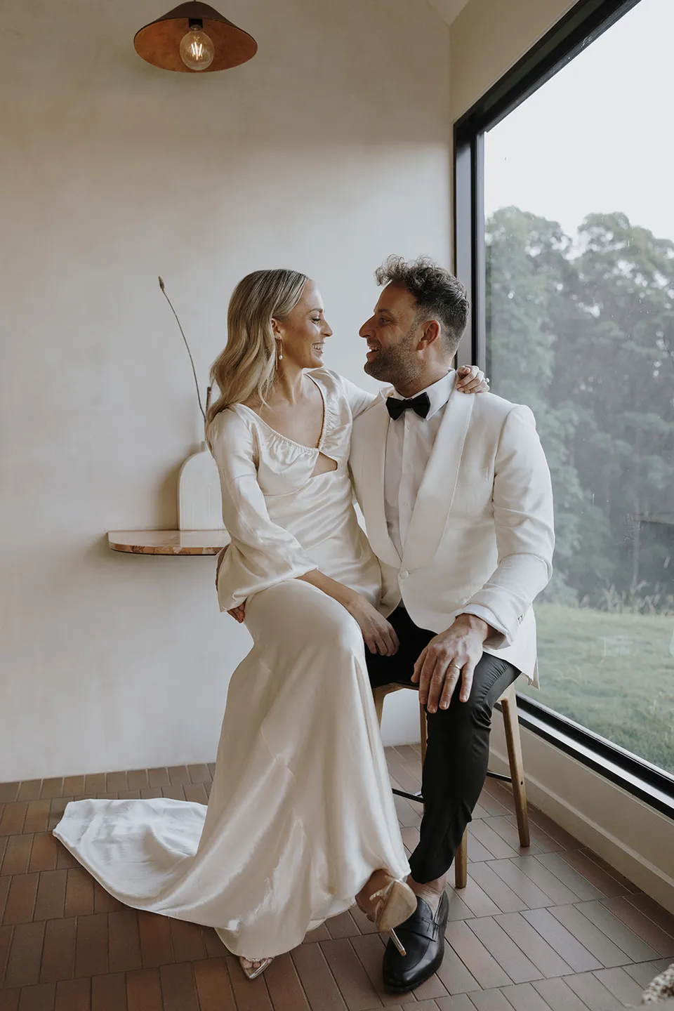Gabrielle and Greg looking at each other on the window seat beside a broad view of the hinterland, at Roundhill Retreat in Obi Obi, Sunshine Coast Hinterland, Queensland, for an Elopement Collective elopement. Photo by George Bowden.