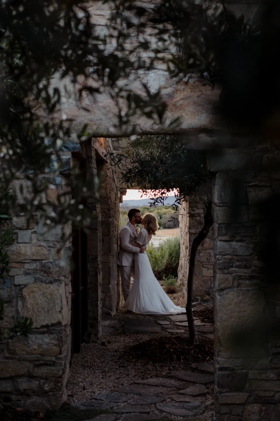 A view through the shaded courtyard towards the couple in a stone doorway, at Thalia Haven on the East Coast of Tasmania, Australia, for an Elopement Collective elopement. Photograph by Joey Willis.