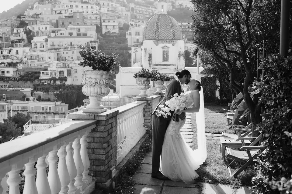Black and white photograph of Skyler and Zach embracing beside the terrace balustrade, with her Flora Garden - Positano bouquet and the hillside town behind them, at Hotel Buca di Bacco in Positano, Amalfi Coast, Italy, for an Elopement Collective elopement. Photograph by Jason Corroto.