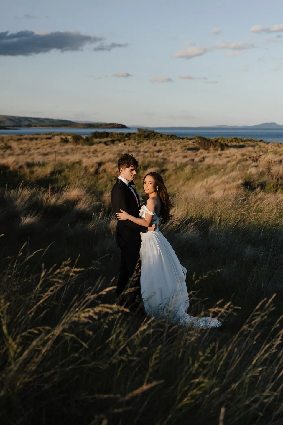 Clara and Harry embracing among tall coastal grasses in warm evening light, with the bay beyond, at Thalia Haven on the East Coast of Tasmania, for an Elopement Collective elopement. Photograph by Bulb Creative.