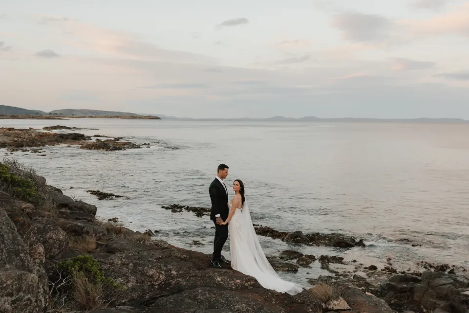 Dani and Anthony facing one another above a rocky cove in soft evening light, at Thalia Haven on Tasmania’s east coast, for an Elopement Collective elopement. Photograph by Bulb Creative.