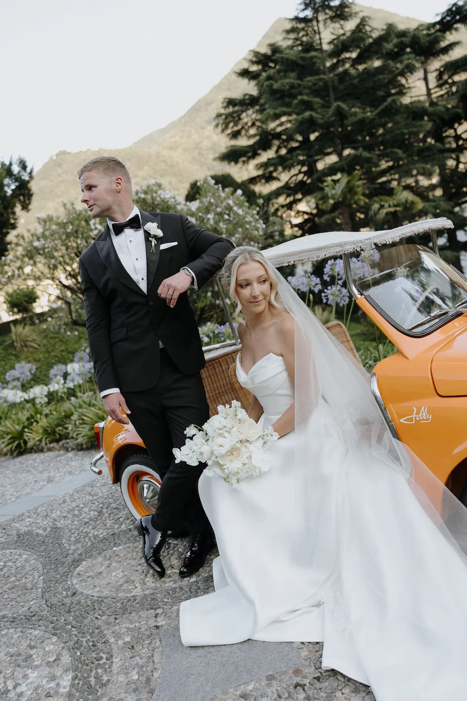 Zoe holding her bouquet by Garden of Love beside Charlton and an orange open-top car on the cobbled driveway, at Passalacqua in Moltrasio, Lake Como, Italy, for an Elopement Collective elopement. Photo by George Bowden.