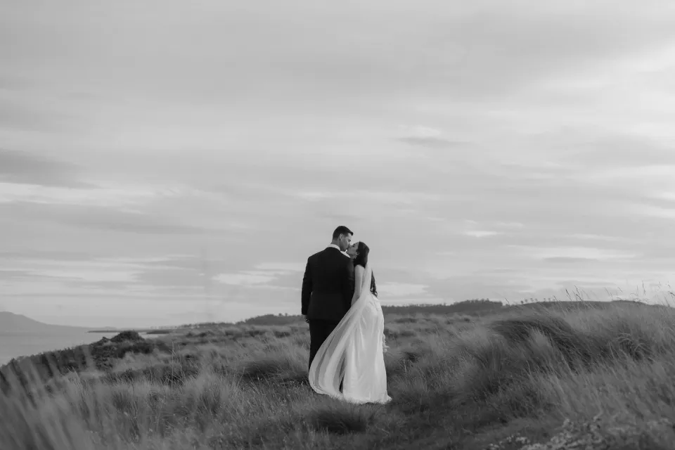 Black and white photograph of Dani and Anthony walking away through coastal grass, with her veil lifting in the breeze, at Thalia Haven on Tasmania’s east coast, for an Elopement Collective elopement. Photograph by Bulb Creative.