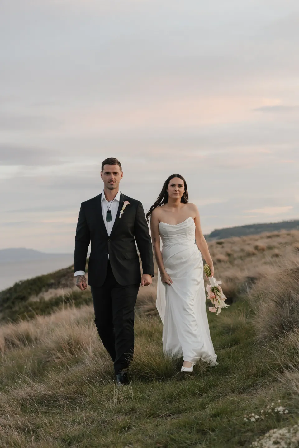 Dani and Anthony holding hands in the coastal grassland at dusk, with her bouquet by The Romantics, at Thalia Haven on Tasmania’s east coast, for an Elopement Collective elopement. Photograph by Bulb Creative.