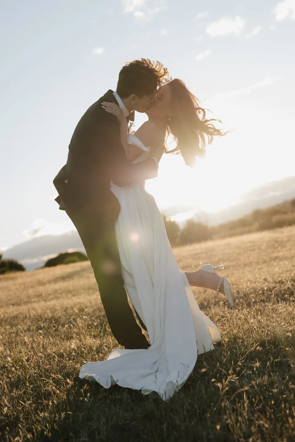 Harry lifting Clara into a kiss, with sunlight behind her windblown hair and flowing white dress, at Thalia Haven on the East Coast of Tasmania, for an Elopement Collective elopement. Photograph by Bulb Creative.