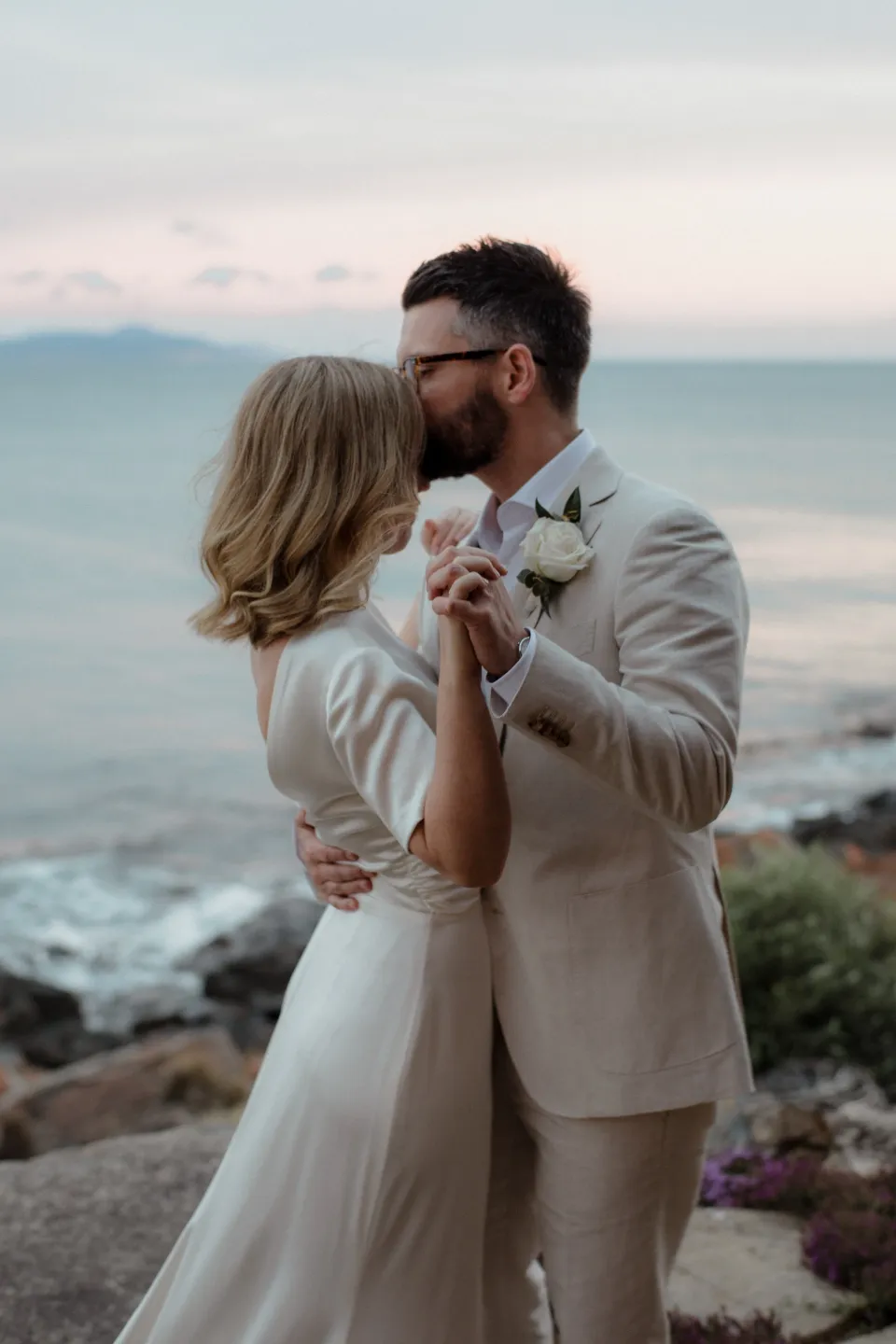Jack kissing Jemmah on the terrace beneath the pastel evening sky, at Thalia Haven on the East Coast of Tasmania, Australia, for an Elopement Collective elopement. Photograph by Joey Willis.
