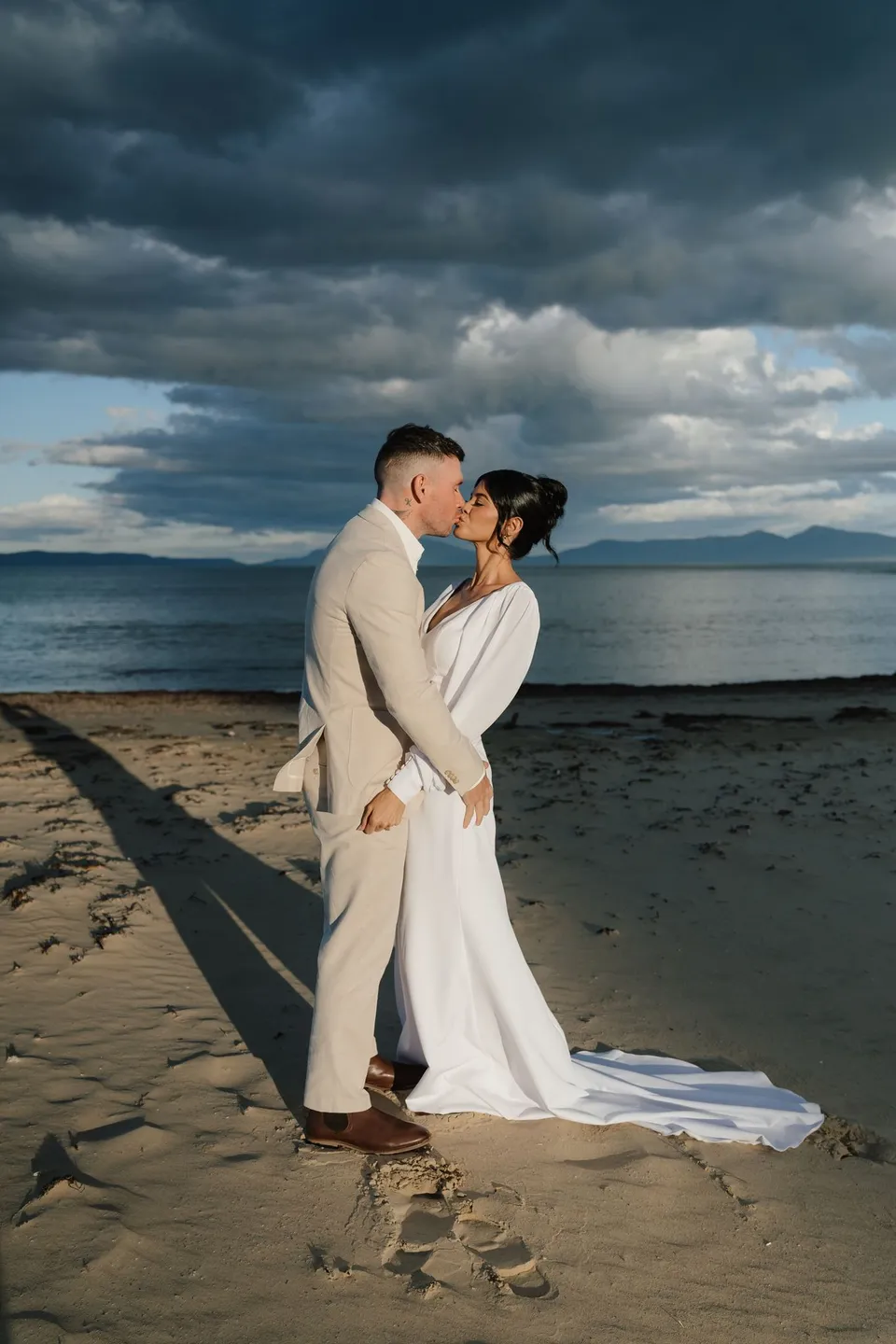 Renee and Mitch kissing on the beach in warm light beneath a dark, cloud-filled sky. At Piermont Retreat on Tasmania’s east coast, for an Elopement Collective elopement. Photograph by Bulb Creative.