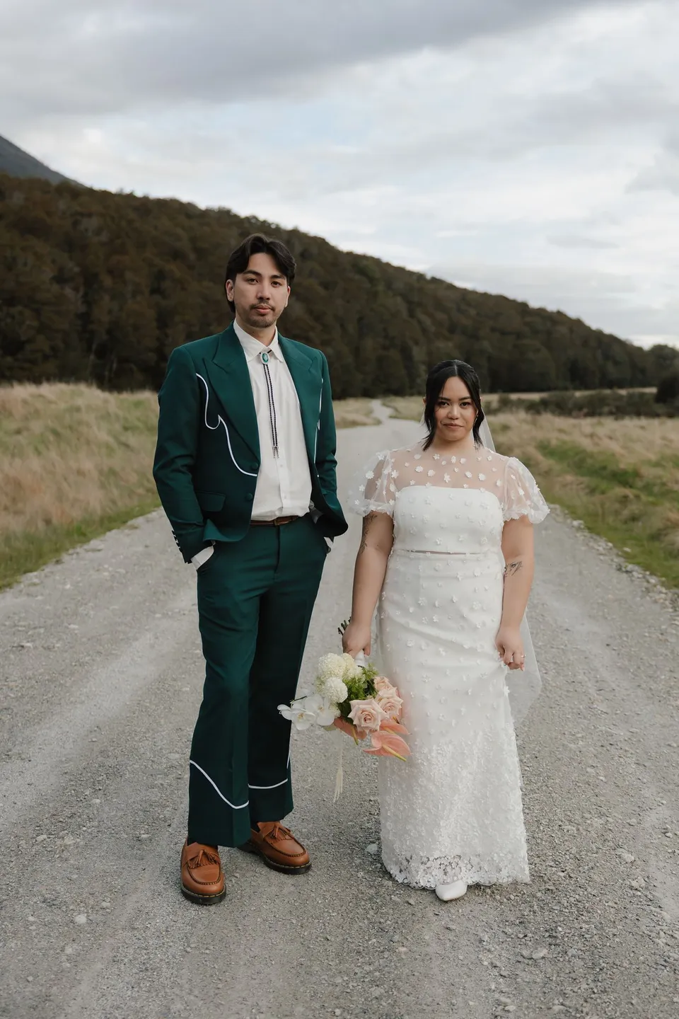 Jaeda and Adrian standing together on a gravel track between green hills, with her Orange Blossom Designs bouquet, at Moke Lake, Queenstown, New Zealand, for an Elopement Collective Queenstown elopement. Photograph by Bulb Creative.