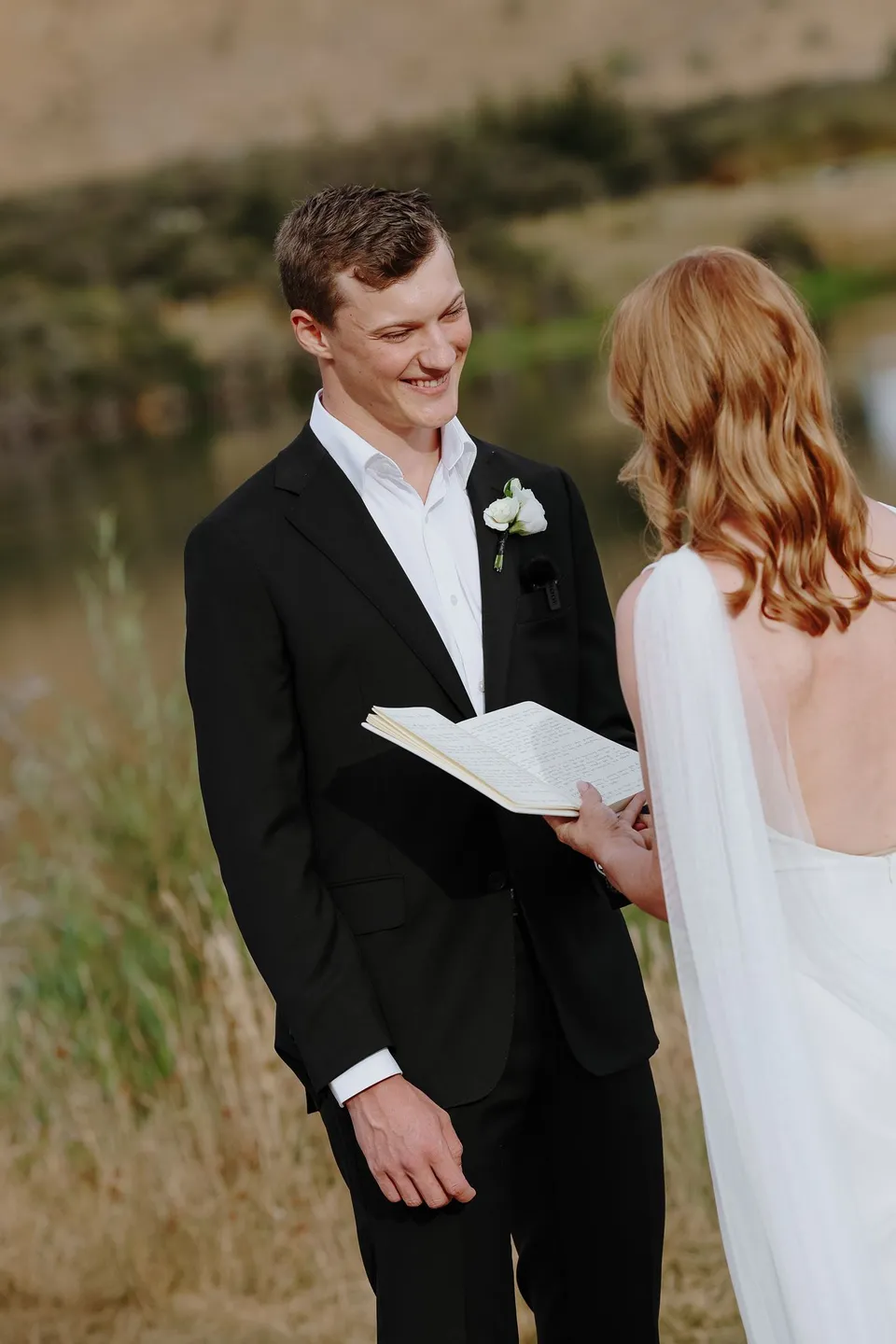Hamish smiling at Neve as she reads from her vow pages beside the lake, at Moke Lake in Queenstown, New Zealand, for an Elopement Collective Queenstown elopement. Photo by George Bowden.