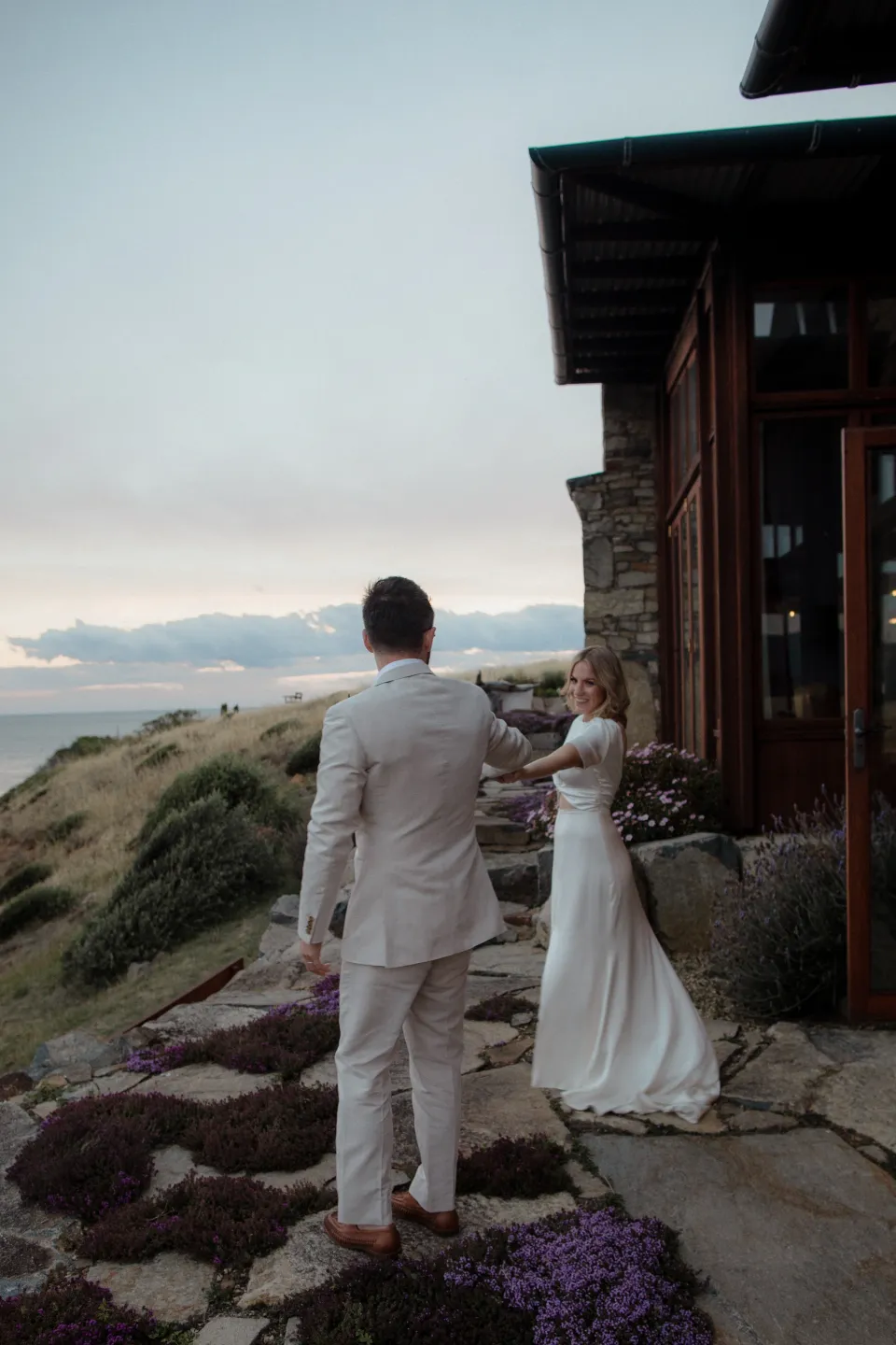 Rear view of the couple beside the house and garden in the evening light, at Thalia Haven on the East Coast of Tasmania, Australia, for an Elopement Collective elopement. Photograph by Joey Willis.