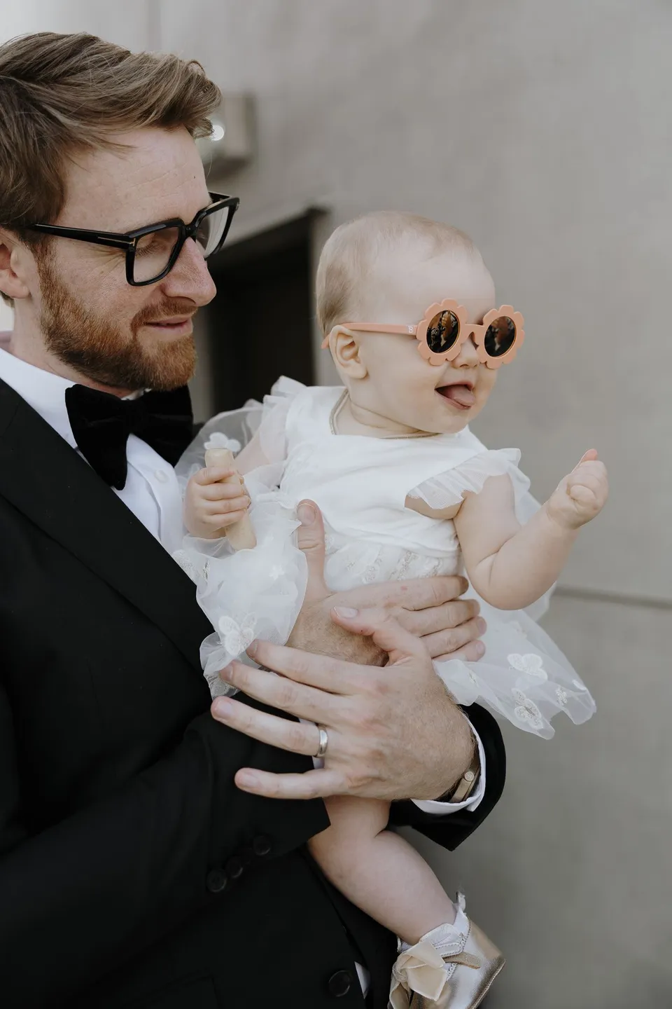 Adam holding a baby wearing round sunglasses and a white outfit, at The Calile Hotel in Fortitude Valley, Brisbane, for an Elopement Collective elopement. Photo by George Bowden.