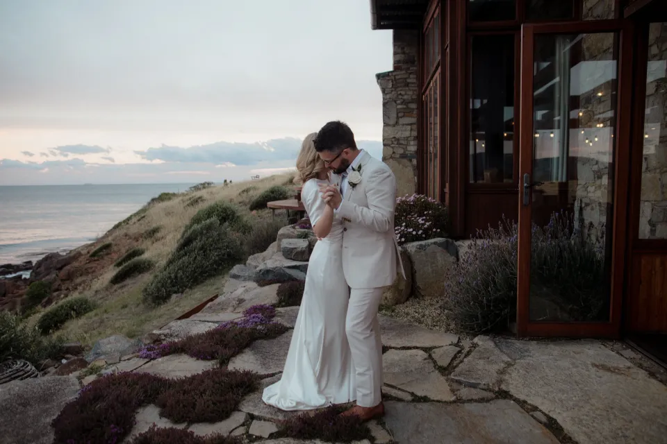Jemmah and Jack embracing beside the stone house with the bay beyond, at Thalia Haven on the East Coast of Tasmania, Australia, for an Elopement Collective elopement. Photograph by Joey Willis.