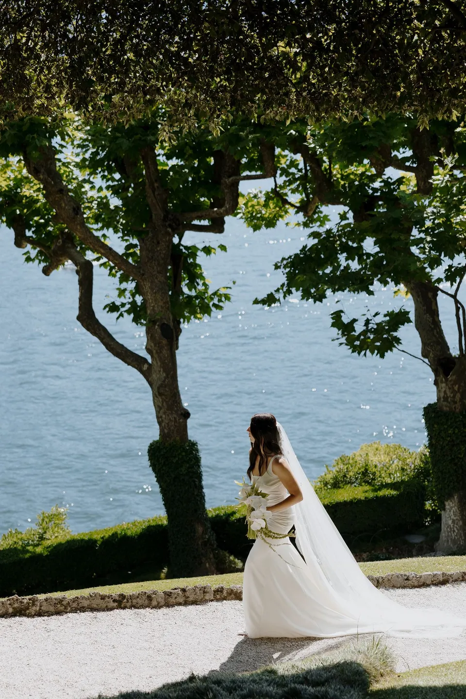 Lyndal walking along a garden path at Villa del Balbianello with her white dress and veil trailing behind her above the lake, at Lake Como in Lombardy, Italy, for an Elopement Collective elopement. Photograph by George Bowden.